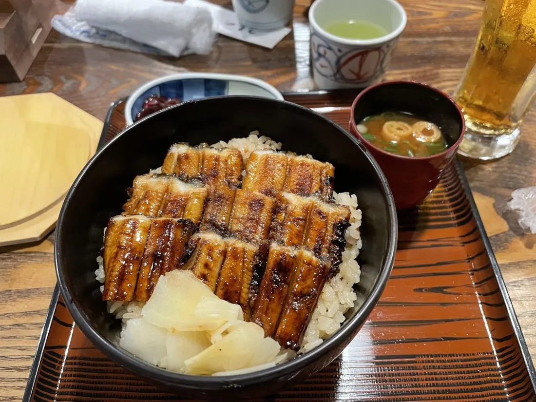 A bowl of grilled eel over rice with miso soup and drinks on a wooden table